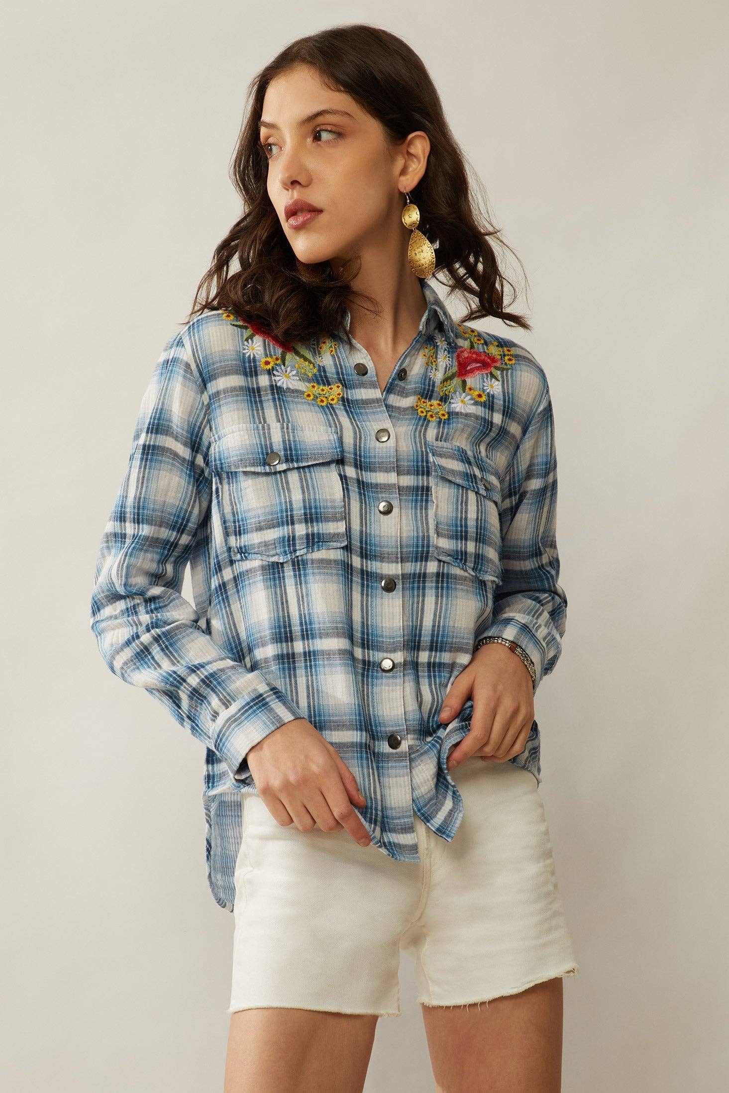 Woman wearing a blue plaid shirt with floral embroidery and white shorts on a plain background